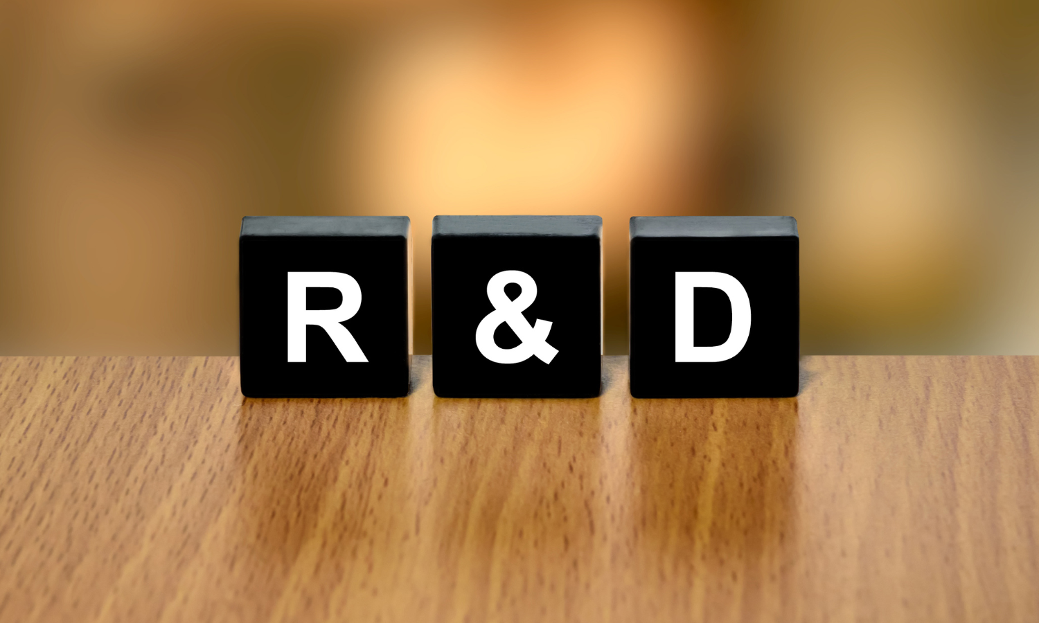 R&D is in three black letter blocks on a table with a blurred gold background